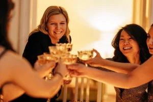 Young attractive women clink glasses of champagne. Charming multiethnic female friends in fancy dresses raise a toast at a bachelorette party. A fun party at home in the living room.