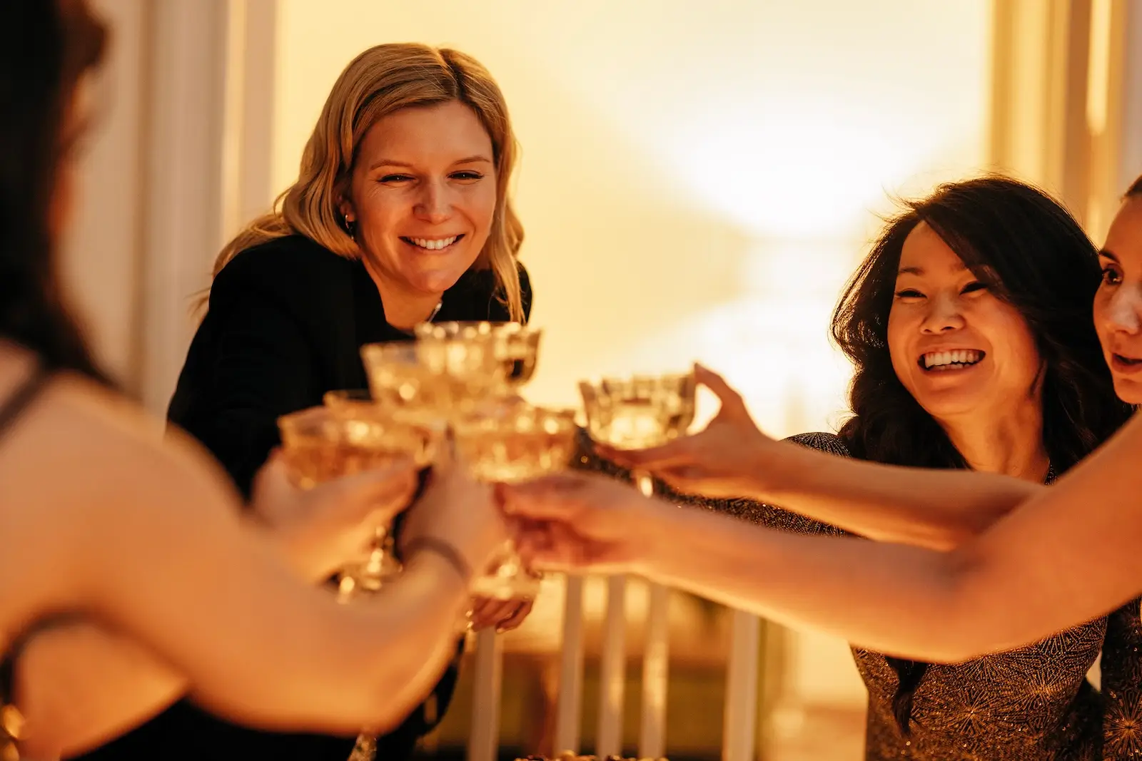 Young attractive women clink glasses of champagne. Charming multiethnic female friends in fancy dresses raise a toast at a bachelorette party. A fun party at home in the living room.